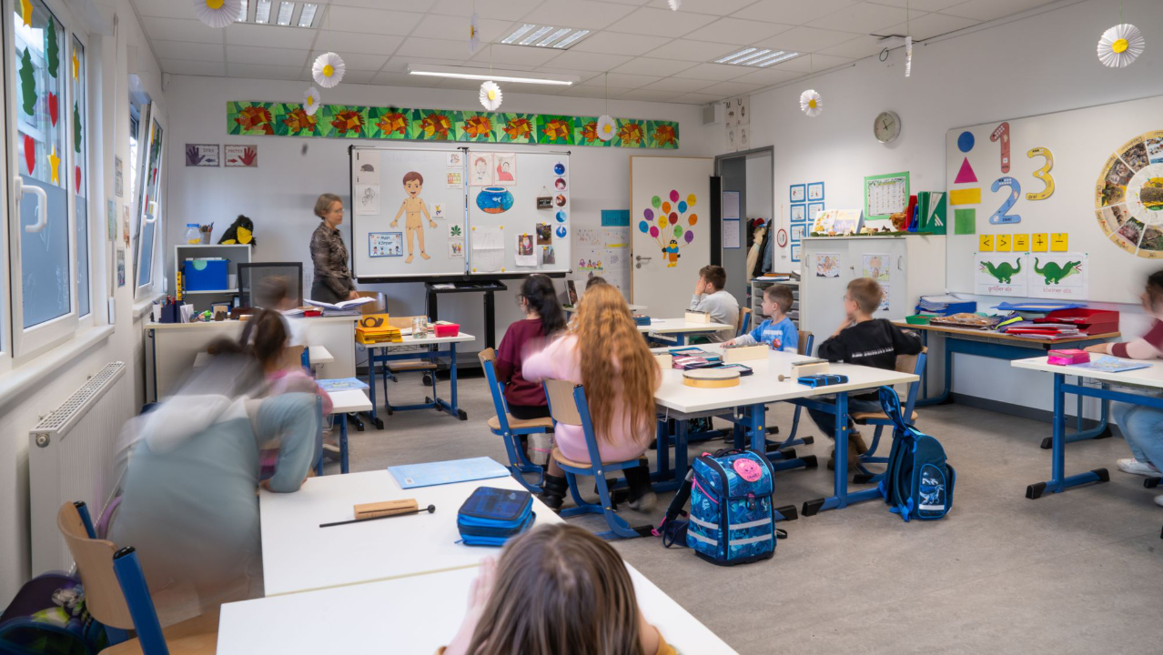 Eine Lehrerin steht vorne am Whiteboard und mehrere Schüler sitzen an Tischen im Klassenraum
