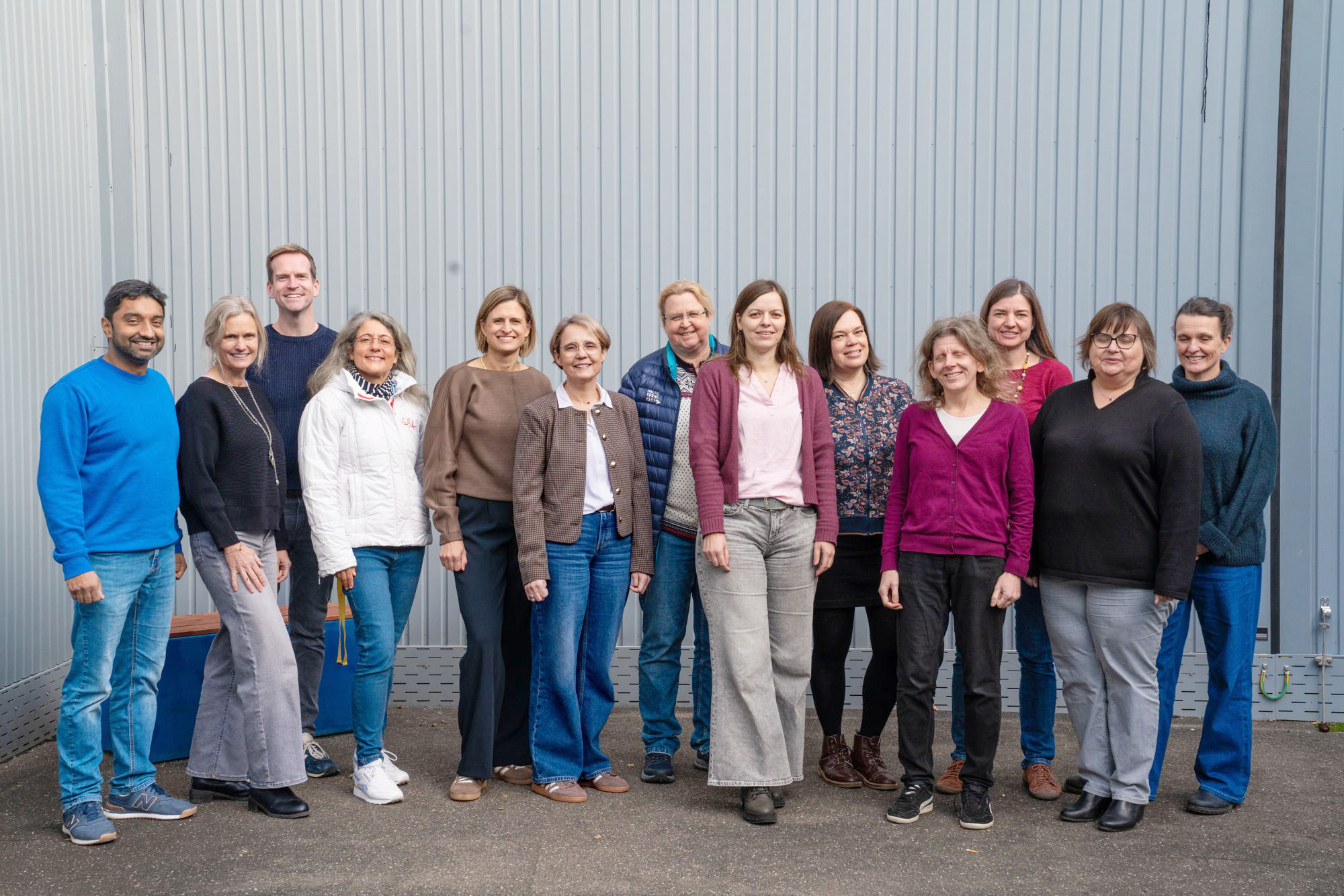 12 Lehrerinnen und Lehrer stehen vor einer grauen Wand und schauen lächelnd in die Kamera