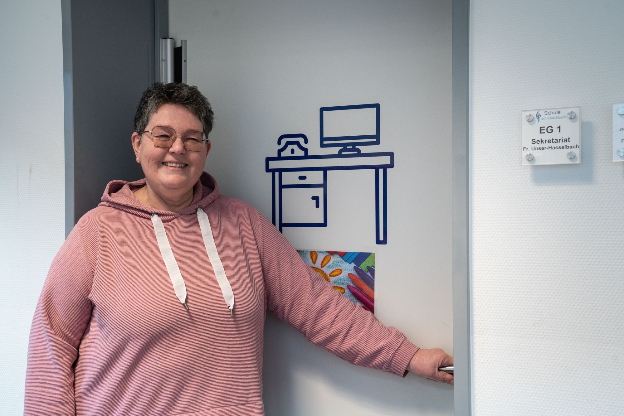 Die Schulsekretaerin Frau Unser-Hasselbach lächelt an einer Tür und neben ihr hängt ein Schild für Sekretariat
