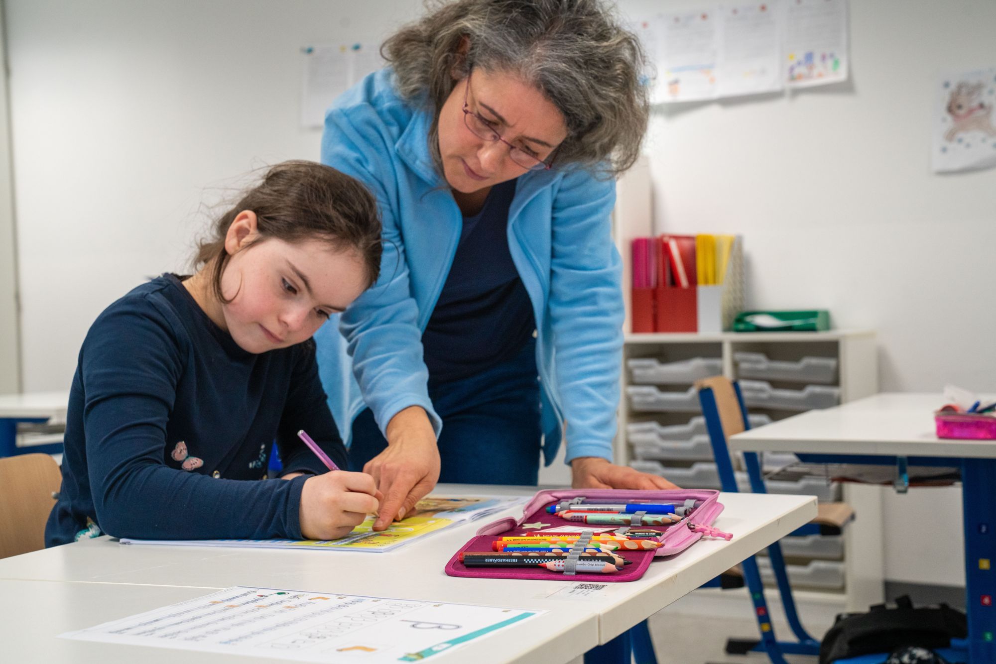 Ein Mädchen schreibt im Heft und eine Lehrerin beugt sich freundlich dazu am Tisch