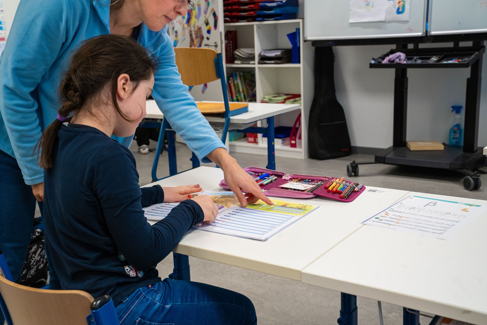 Eine Lehrerin zeigt einem Mädchen am Tisch die Aufgabe und Stifte liegen offen daneben