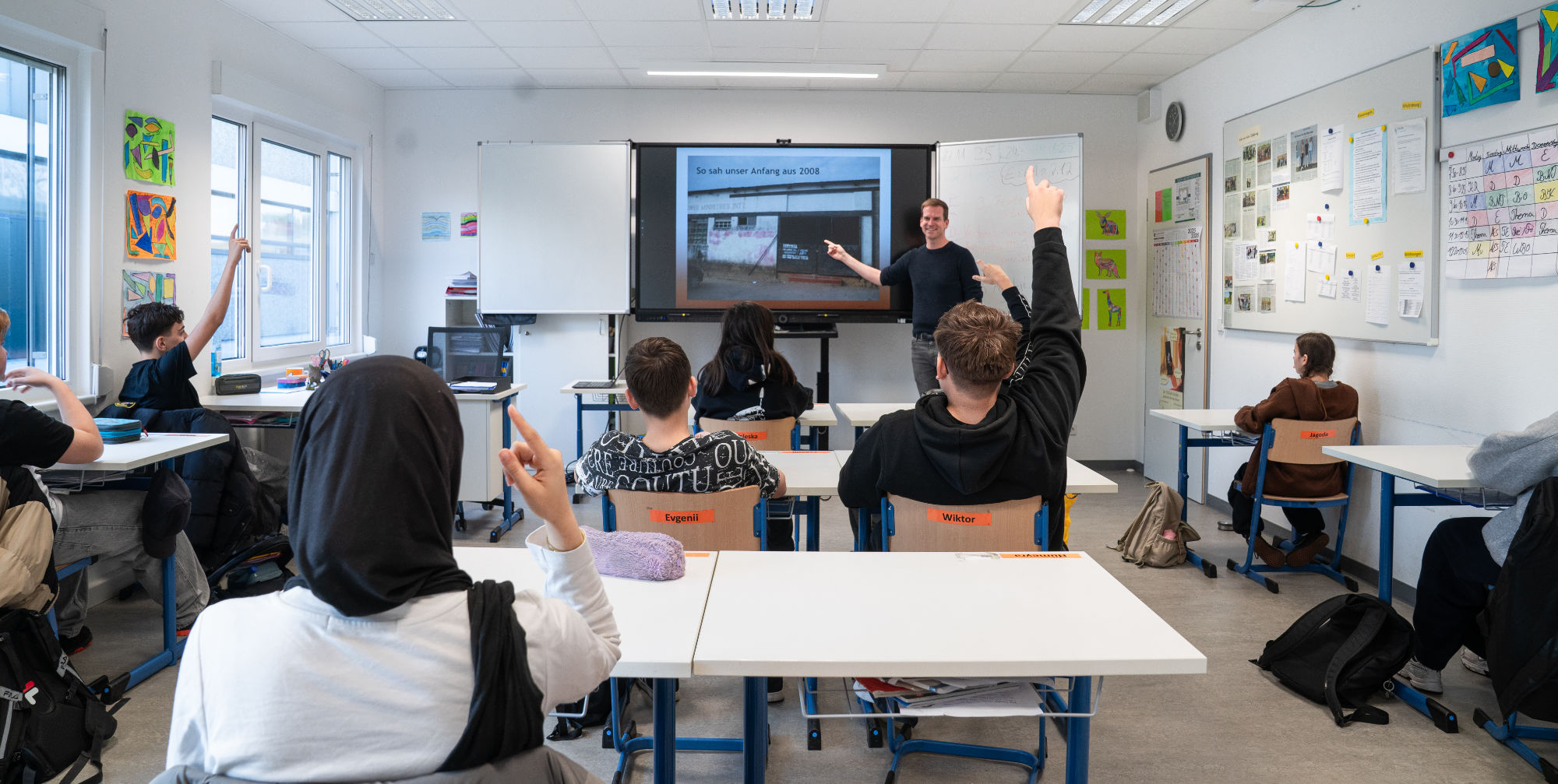 Schüler einer Klasse melden sich zu einem Projekt in Afrika zur Beschaffung von Brillen für Schüler Der Lehrer zeigt auf die Videowand
