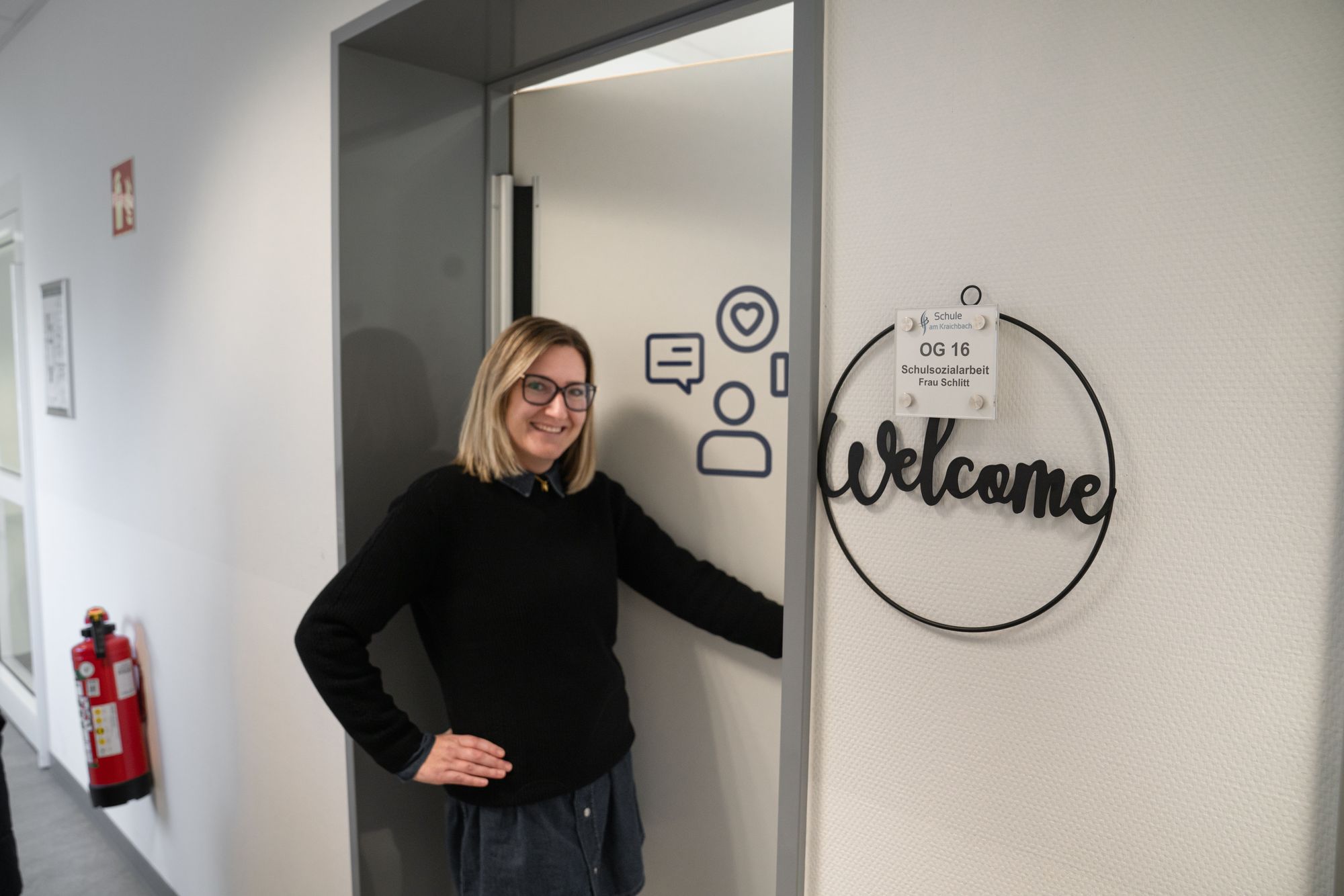 Schulsozialarbeiterin Frau Schlitt steht in der Tür und lächelt neben einem Welcome Schild und einem Türschild